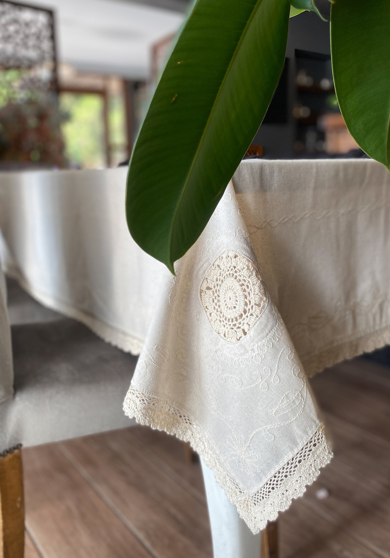 May include: A white linen tablecloth with delicate crocheted trim and a circular design in the center. The tablecloth is draped over a table and chair, with a green leaf in the background.