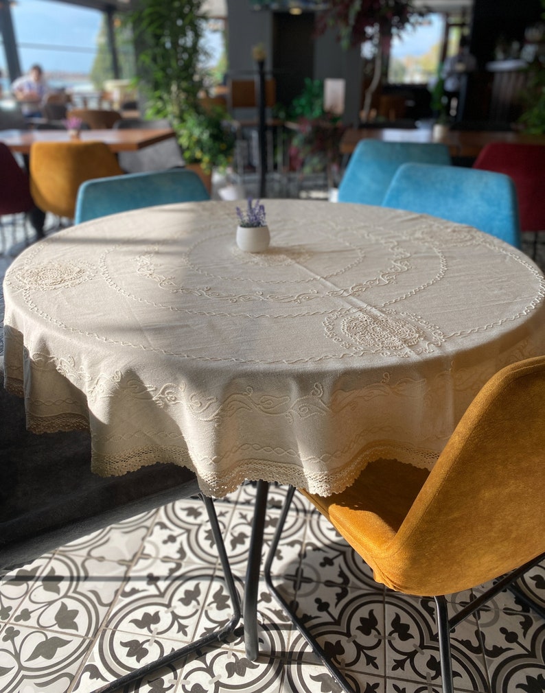 Pu&ograve; includere: Un tavolo rotondo con una tovaglia bianca e un bordo in pizzo. Il tavolo &egrave; apparecchiato con un piccolo vaso bianco con fiori di lavanda. Ci sono quattro sedie intorno al tavolo, due blu e due gialle.