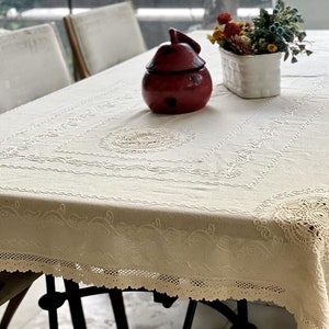 May include: A white tablecloth with delicate lace trim and embroidered details. A red ceramic jar and a white planter with dried flowers are on the table.