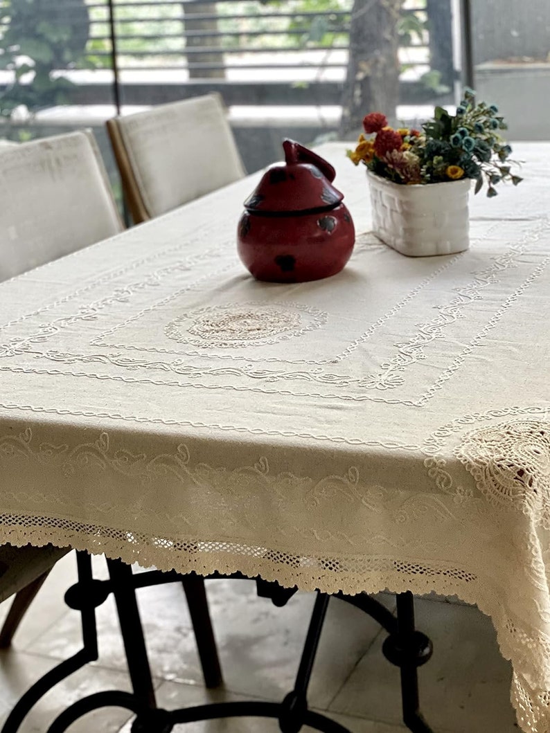 May include: A white tablecloth with delicate lace trim and embroidered details. A red ceramic jar and a white planter with dried flowers are on the table.