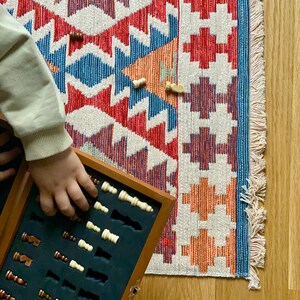 May include: A wooden chessboard box with a black felt interior and a set of white and brown chess pieces. The box is open and the pieces are arranged in rows. The box is sitting on a colorful rug with a geometric pattern.