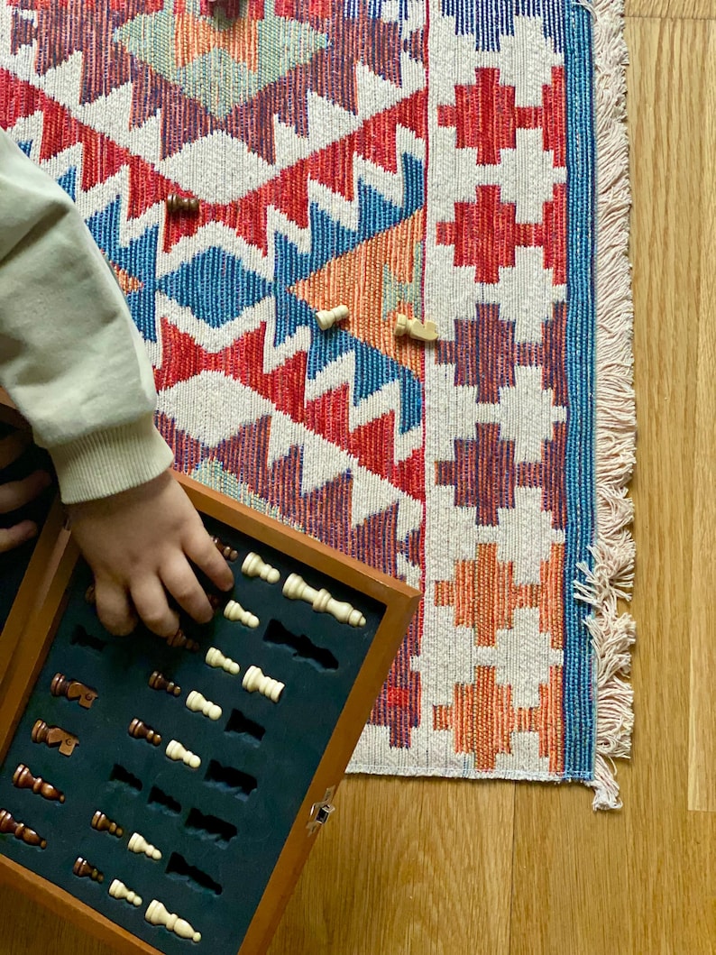 May include: A wooden chessboard box with a black felt interior and a set of white and brown chess pieces. The box is open and the pieces are arranged in rows. The box is sitting on a colorful rug with a geometric pattern.