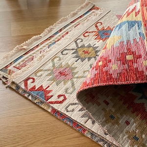 May include: A folded rug with a white background and a colorful geometric pattern in red, blue, and yellow. The rug has a fringe border.