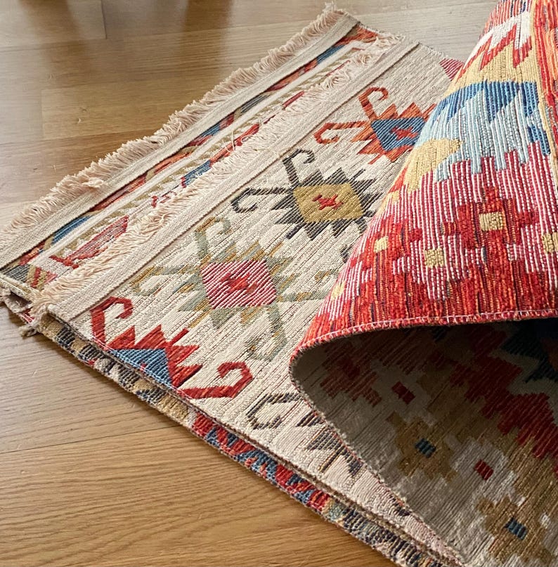May include: A folded rug with a white background and a colorful geometric pattern in red, blue, and yellow. The rug has a fringe border.