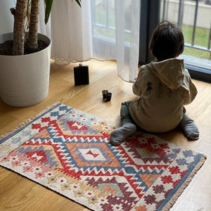 May include: A colorful geometric patterned rug with fringe on the edges. A child sits on the rug looking out a window.