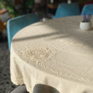 Pu&ograve; includere: Un primo piano di una tovaglia bianca con un delicato bordo di pizzo e dettagli ricamati. La tovaglia &egrave; drappeggiata su un tavolo rotondo, con un piccolo vaso bianco con una pianta verde sul tavolo.