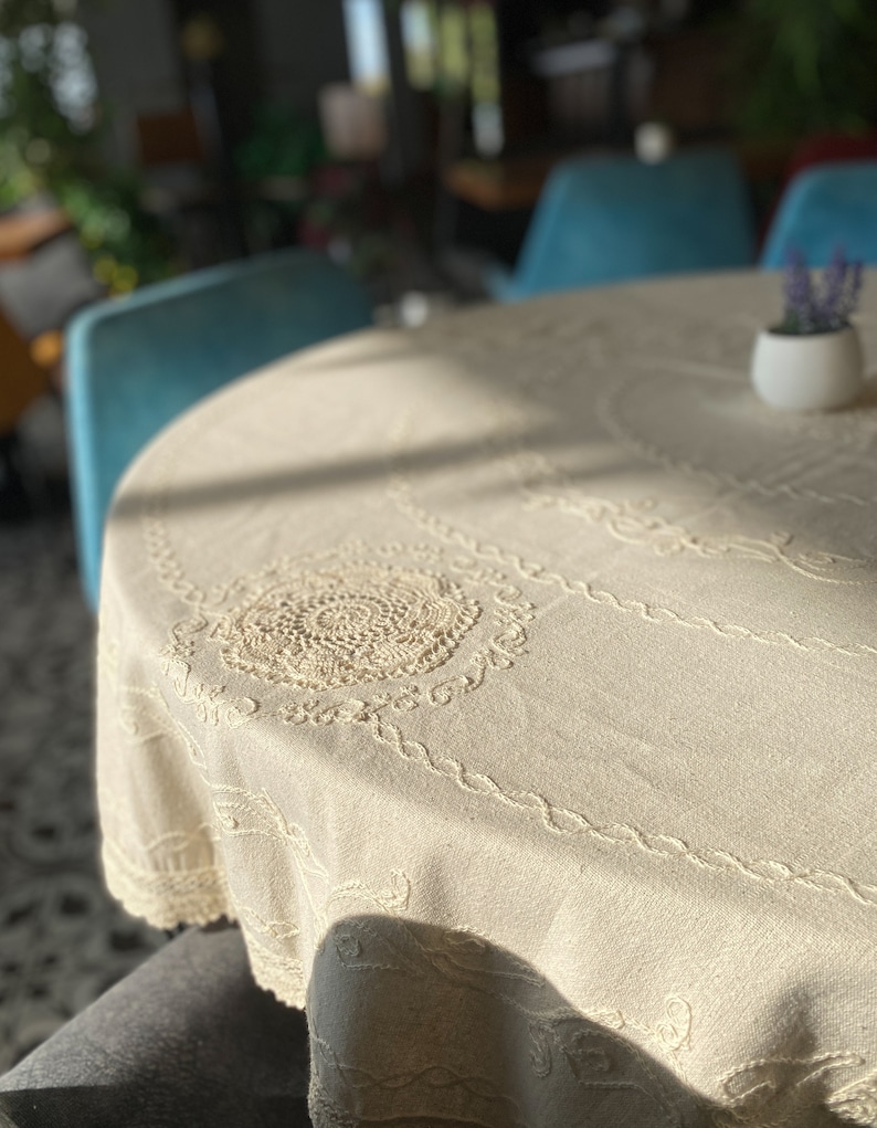 Pu&ograve; includere: Un primo piano di una tovaglia bianca con un delicato bordo di pizzo e dettagli ricamati. La tovaglia &egrave; drappeggiata su un tavolo rotondo, con un piccolo vaso bianco con una pianta verde sul tavolo.