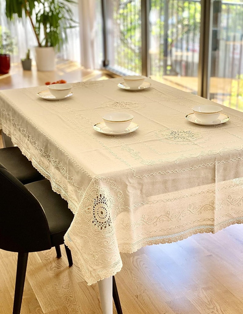 May include: A white tablecloth with delicate lace trim and embroidered details. Four white bowls with gold rims are placed on the tablecloth, creating a simple and elegant table setting.
