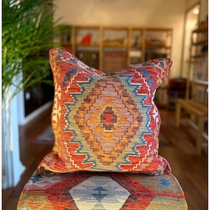 Puede incluir: Un cojín decorativo con un vibrante patrón geométrico en rojo, naranja, azul y amarillo. El cojín está colocado sobre un banco con un patrón similar. El diseño presenta formas de diamantes y zigzags, creando una apariencia colorida y texturizada.