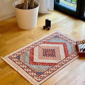Puede incluir: Una alfombra pequeña y rectangular con un patrón geométrico en tonos rojos, azules y crema. La alfombra tiene un borde de flecos y está colocada sobre un suelo de madera.