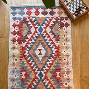 May include: A colorful rug with a geometric pattern in shades of red, blue, orange, and beige. The rug has a fringe border and is made of woven fabric.