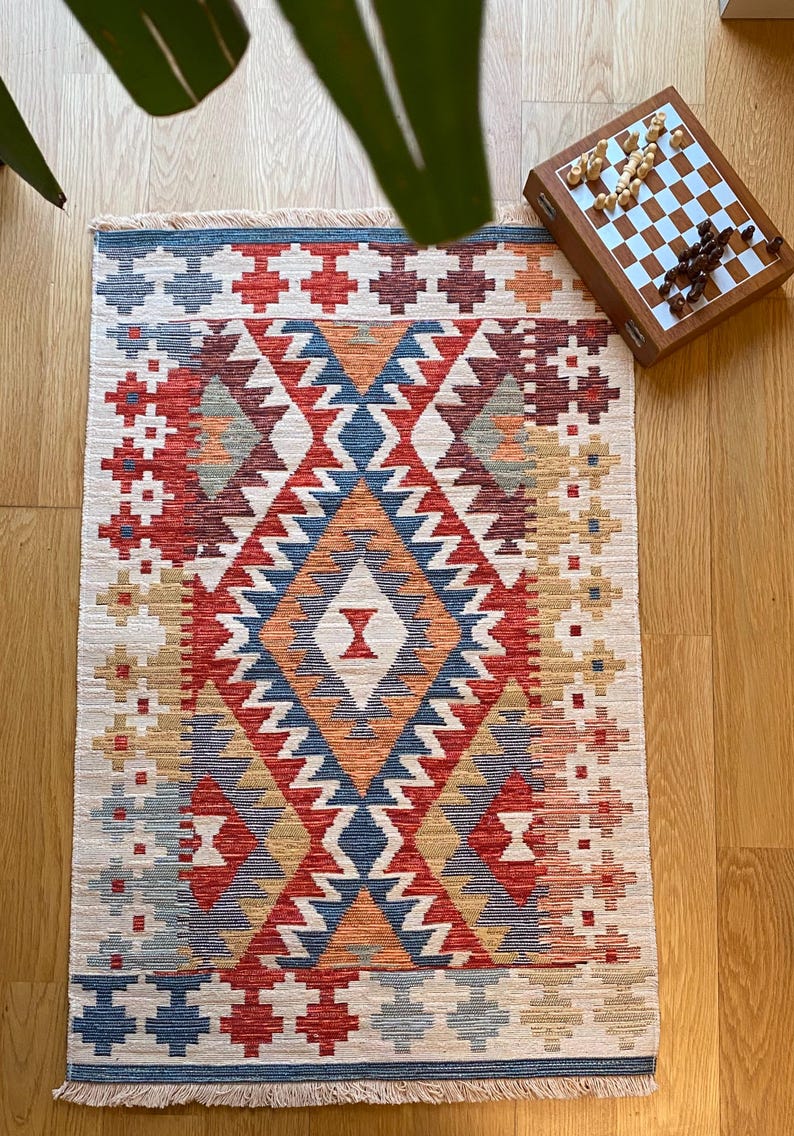 May include: A colorful rug with a geometric pattern in shades of red, blue, orange, and beige. The rug has a fringe border and is made of woven fabric.