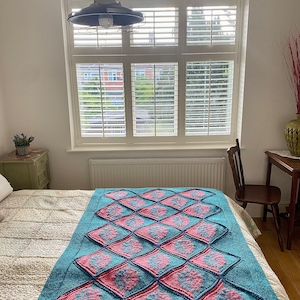 K&ouml;nnte beinhalten: Eine tealblaue gestrickte Wolldecke mit einem Rautenmuster, das mit rosa und blauen Blumenmustern verziert ist. Die Decke liegt auf einem Bett mit einer wei&szlig;en Bettw&auml;sche mit Blumenmuster.