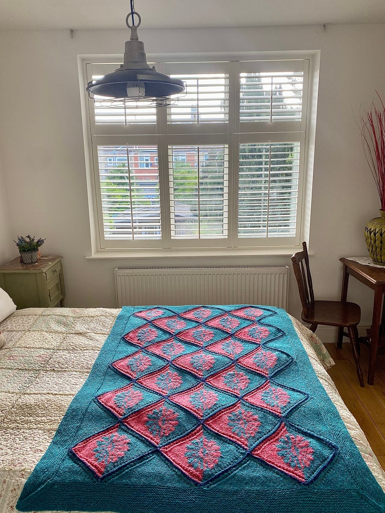 K&ouml;nnte beinhalten: Eine tealblaue gestrickte Wolldecke mit einem Rautenmuster, das mit rosa und blauen Blumenmustern verziert ist. Die Decke liegt auf einem Bett mit einer wei&szlig;en Bettw&auml;sche mit Blumenmuster.