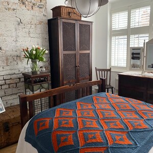 Pode incluir: Uma manta de malha azul e laranja com um padr&atilde;o geom&eacute;trico em uma cama em um quarto com um arm&aacute;rio de madeira e uma parede de tijolo.