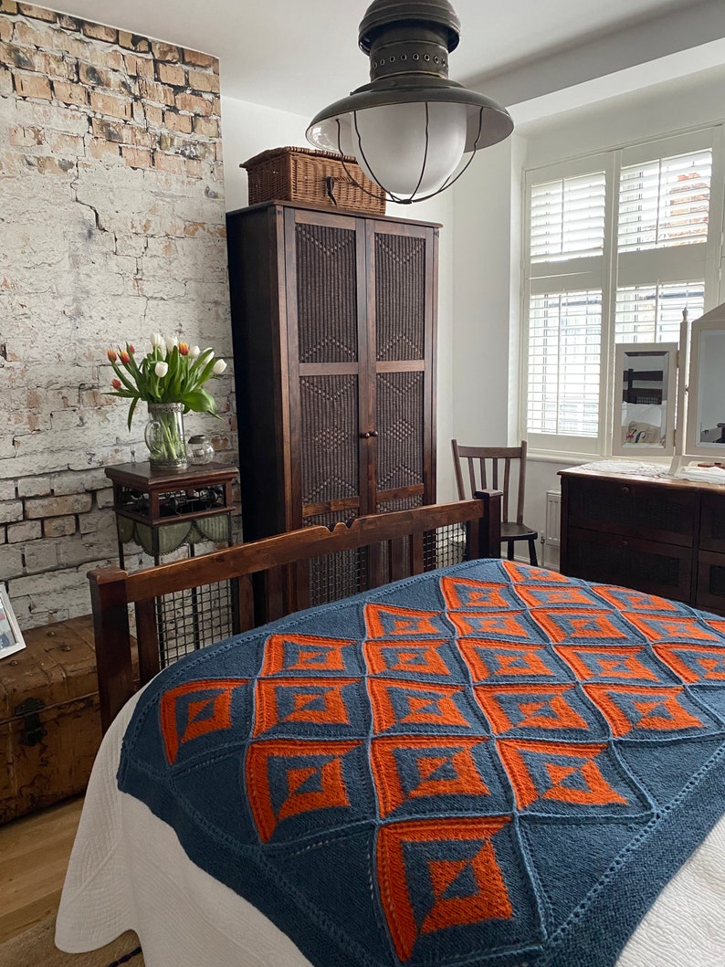Pode incluir: Uma manta de malha azul e laranja com um padr&atilde;o geom&eacute;trico em uma cama em um quarto com um arm&aacute;rio de madeira e uma parede de tijolo.