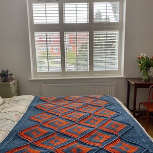 Pode incluir: Uma manta de malha azul e laranja com um padr&atilde;o geom&eacute;trico. A manta est&aacute; dobrada e colocada em uma cama com roupa de cama branca.