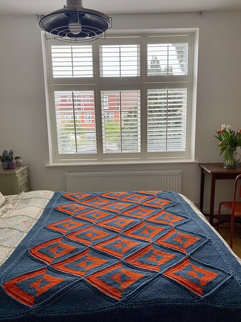Pode incluir: Uma manta de malha azul e laranja com um padr&atilde;o geom&eacute;trico. A manta est&aacute; dobrada e colocada em uma cama com roupa de cama branca.