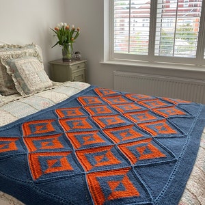 Pode incluir: Uma manta de malha azul e laranja com um padr&atilde;o geom&eacute;trico. A manta est&aacute; em uma cama com uma colcha branca com padr&atilde;o floral.