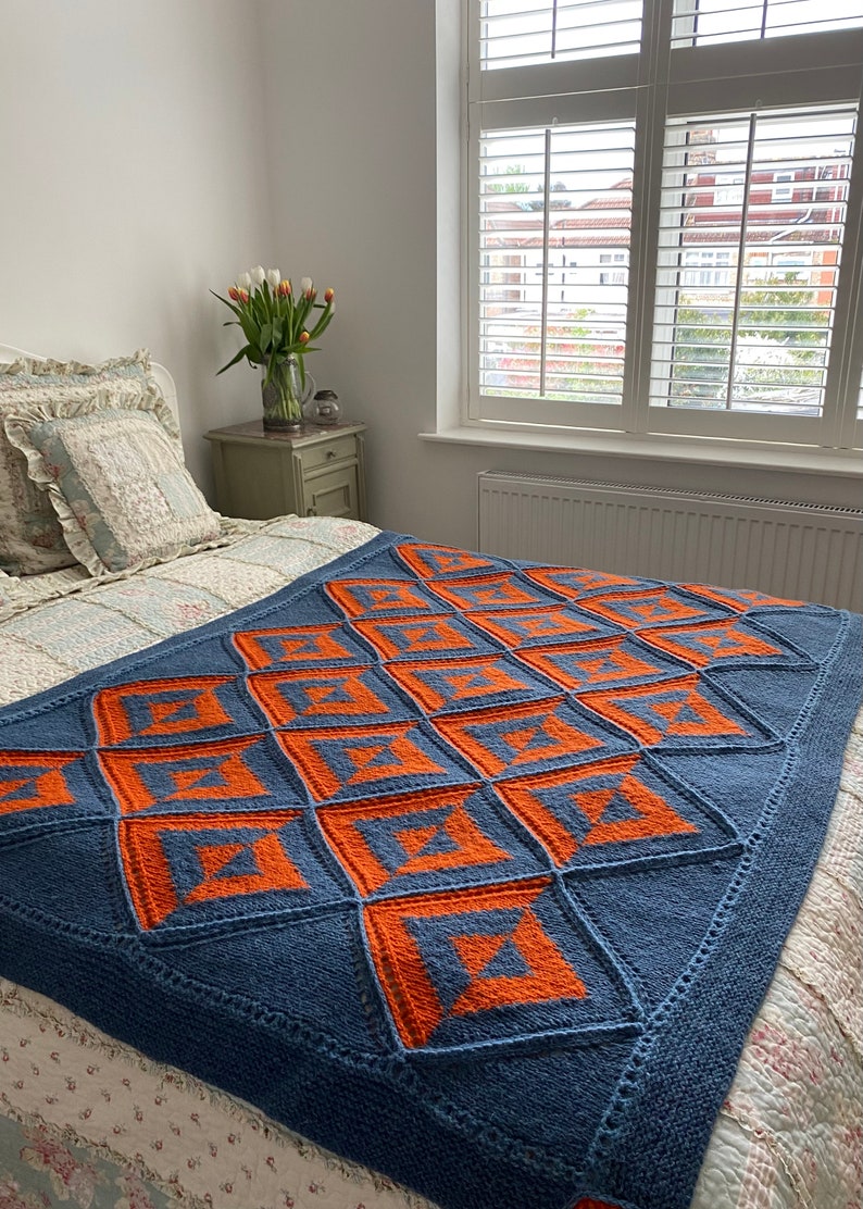 Pode incluir: Uma manta de malha azul e laranja com um padr&atilde;o geom&eacute;trico. A manta est&aacute; em uma cama com uma colcha branca com padr&atilde;o floral.