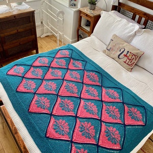 K&ouml;nnte beinhalten: Eine tealfarbene und rosafarbene geh&auml;kelte Tagesdecke mit einem Rautenmuster. Die Decke liegt auf einem Bett mit wei&szlig;en Bettw&auml;sche und einem Kissen mit dem Wort "LOVE" darauf.