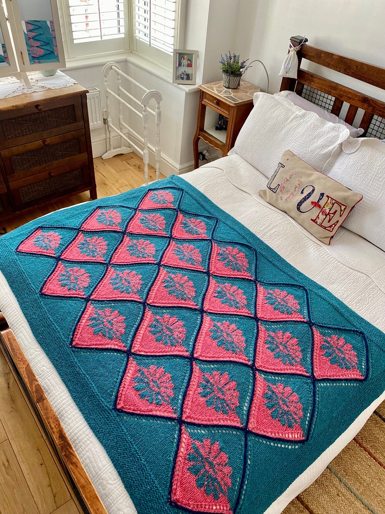 K&ouml;nnte beinhalten: Eine tealfarbene und rosafarbene geh&auml;kelte Tagesdecke mit einem Rautenmuster. Die Decke liegt auf einem Bett mit wei&szlig;en Bettw&auml;sche und einem Kissen mit dem Wort "LOVE" darauf.