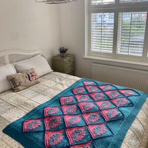 K&ouml;nnte beinhalten: Eine tealfarbene und rosafarbene gestrickte Wolldecke mit geometrischem Muster. Die Decke liegt auf einem Bett mit einem wei&szlig;en Kopfteil und einer wei&szlig;en Bettw&auml;sche mit Blumenmuster.