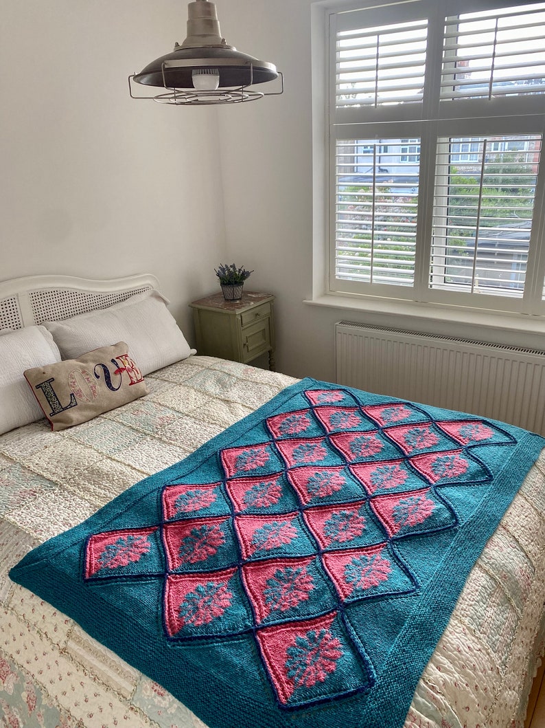 K&ouml;nnte beinhalten: Eine tealfarbene und rosafarbene gestrickte Wolldecke mit geometrischem Muster. Die Decke liegt auf einem Bett mit einem wei&szlig;en Kopfteil und einer wei&szlig;en Bettw&auml;sche mit Blumenmuster.