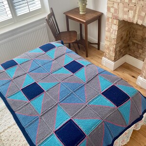 May include: A knitted throw blanket with a geometric pattern in shades of blue, grey, and pink. The blanket is on a bed with a white floral patterned bedspread.