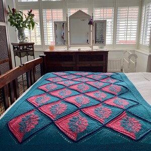 K&ouml;nnte beinhalten: Eine tealblaue gestrickte Wolldecke mit einem geometrischen Muster in Pink und Teal. Die Decke ist gefaltet und auf einem Bett drapiert.