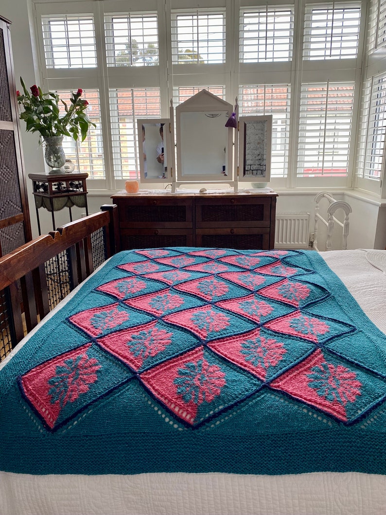 K&ouml;nnte beinhalten: Eine tealblaue gestrickte Wolldecke mit einem geometrischen Muster in Pink und Teal. Die Decke ist gefaltet und auf einem Bett drapiert.