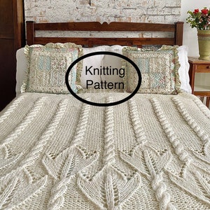 May include: A white knitted throw blanket with a cable knit pattern and a leaf design. The blanket is on a bed with pillows and a wooden headboard.