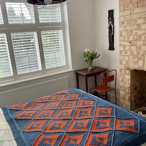 Pode incluir: Uma manta de malha azul e laranja com um padr&atilde;o geom&eacute;trico. A manta est&aacute; em uma cama com uma colcha branca com padr&atilde;o floral.