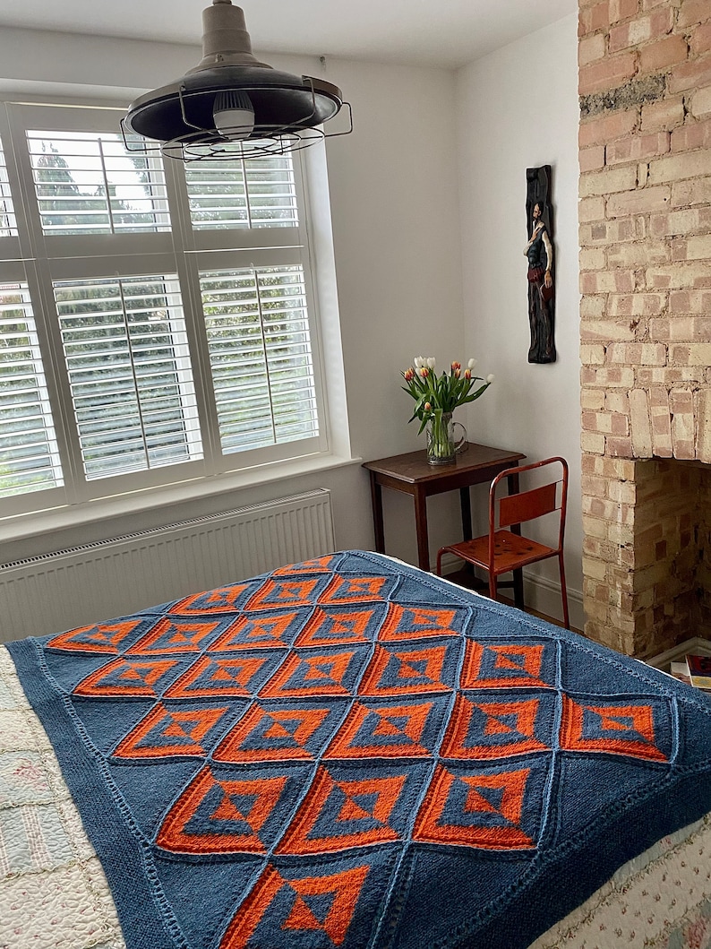 Pode incluir: Uma manta de malha azul e laranja com um padr&atilde;o geom&eacute;trico. A manta est&aacute; em uma cama com uma colcha branca com padr&atilde;o floral.