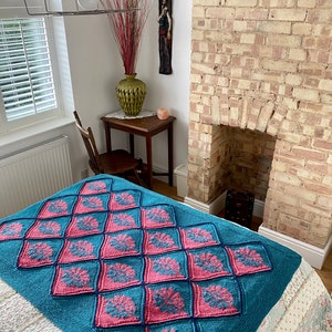 K&ouml;nnte beinhalten: Eine tealblaue und rosafarbene gestrickte Wolldecke mit geometrischem Muster. Die Decke liegt auf einem wei&szlig;en Bett mit Blumenmuster.