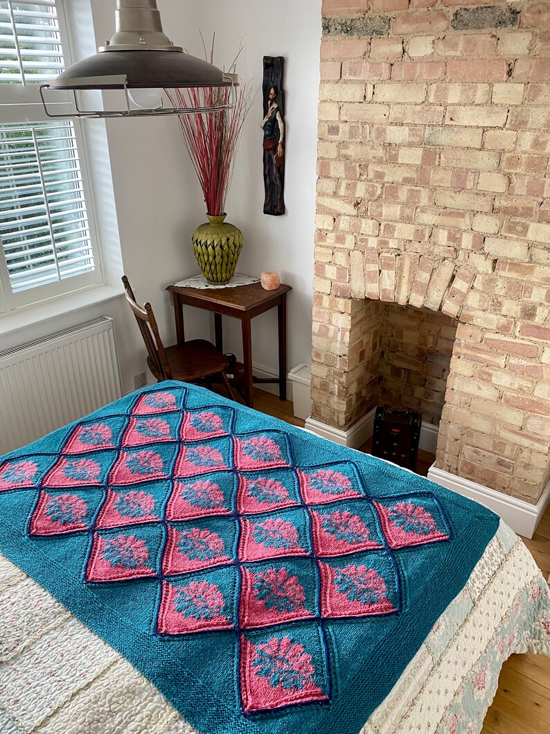 K&ouml;nnte beinhalten: Eine tealblaue und rosafarbene gestrickte Wolldecke mit geometrischem Muster. Die Decke liegt auf einem wei&szlig;en Bett mit Blumenmuster.