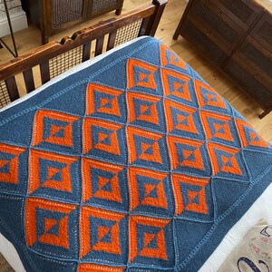 Pode incluir: Uma manta de malha azul e laranja com um padr&atilde;o geom&eacute;trico. A manta est&aacute; em uma cama com um len&ccedil;ol branco e um travesseiro com um padr&atilde;o floral.