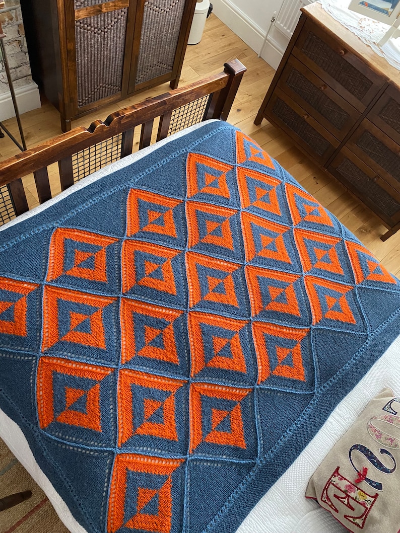Pode incluir: Uma manta de malha azul e laranja com um padr&atilde;o geom&eacute;trico. A manta est&aacute; em uma cama com um len&ccedil;ol branco e um travesseiro com um padr&atilde;o floral.