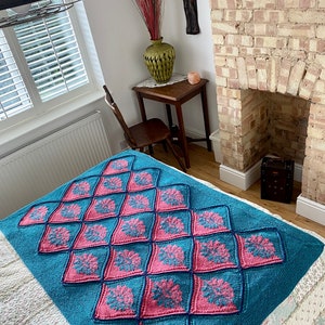K&ouml;nnte beinhalten: Eine tealfarbene und rosafarbene gestrickte Wolldecke mit einem Rautenmuster. Die Decke liegt auf einem Bett mit einer wei&szlig;en Bettw&auml;sche mit Blumenmuster.