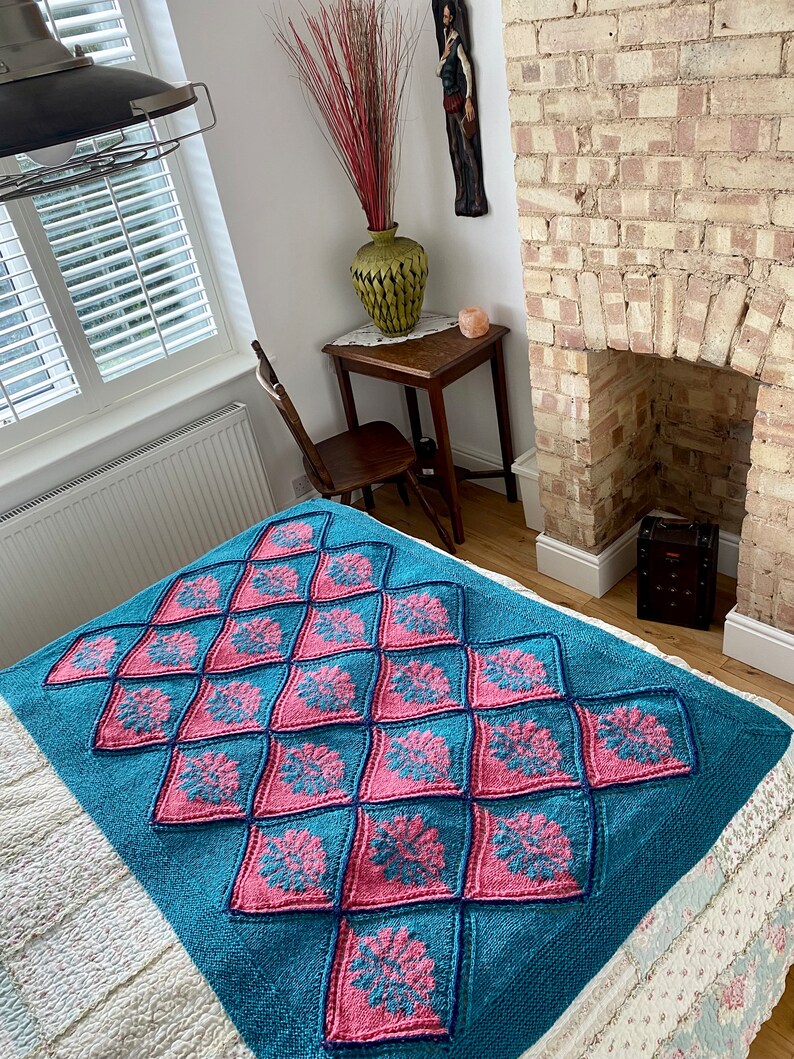 K&ouml;nnte beinhalten: Eine tealfarbene und rosafarbene gestrickte Wolldecke mit einem Rautenmuster. Die Decke liegt auf einem Bett mit einer wei&szlig;en Bettw&auml;sche mit Blumenmuster.