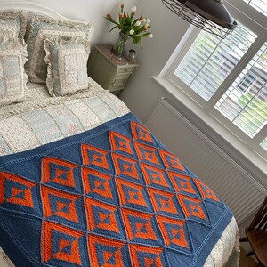 Pode incluir: Uma manta de malha azul e laranja com um padr&atilde;o geom&eacute;trico. A manta est&aacute; em uma cama com roupa de cama floral branca e azul.