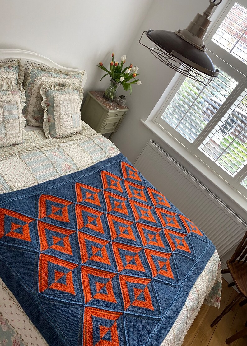 Pode incluir: Uma manta de malha azul e laranja com um padr&atilde;o geom&eacute;trico. A manta est&aacute; em uma cama com roupa de cama floral branca e azul.