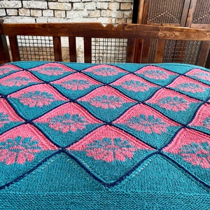 K&ouml;nnte beinhalten: Eine tealfarbene und rosafarbene gestrickte Wolldecke mit einem Rautenmuster und floralem Design.