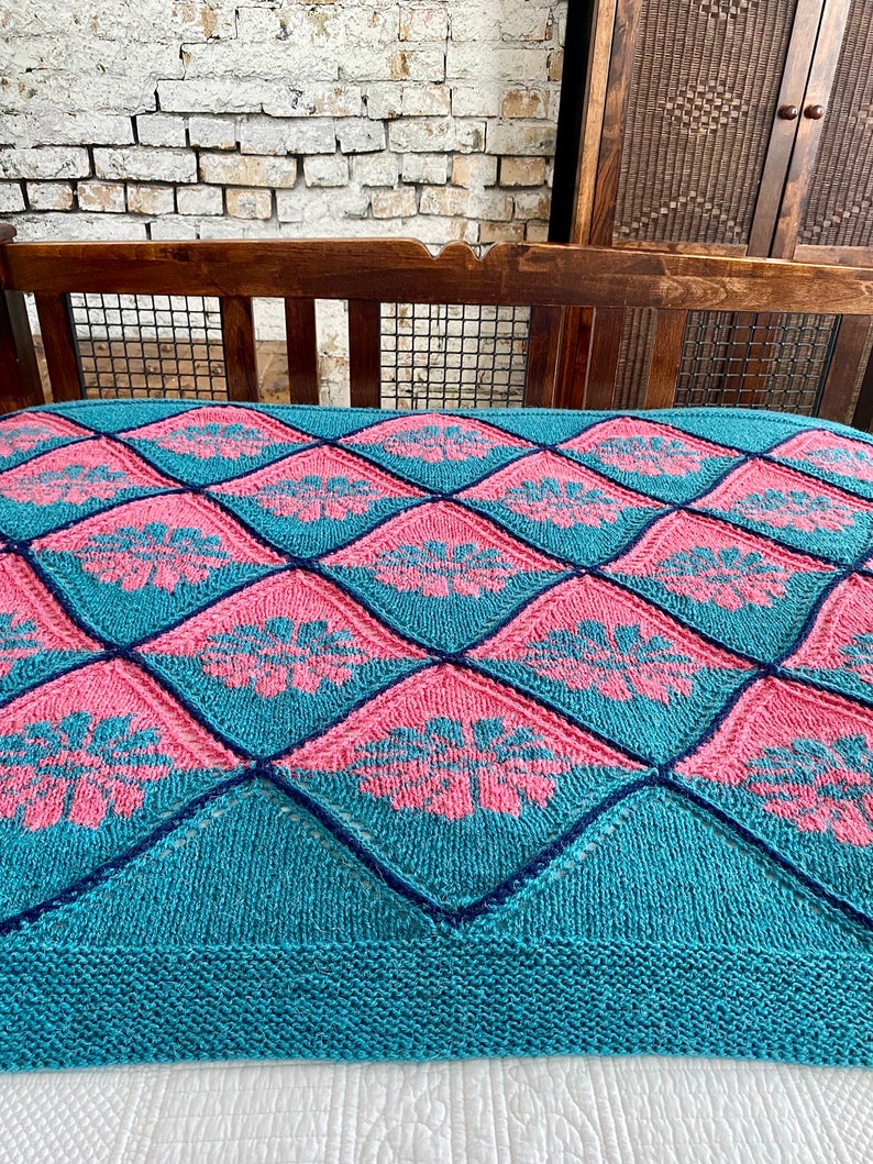 K&ouml;nnte beinhalten: Eine tealfarbene und rosafarbene gestrickte Wolldecke mit einem Rautenmuster und floralem Design.