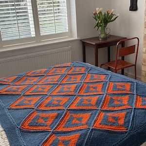 Pode incluir: Uma manta de malha azul e laranja com um padr&atilde;o geom&eacute;trico. A manta est&aacute; em uma cama com uma colcha branca e estampada com flores.