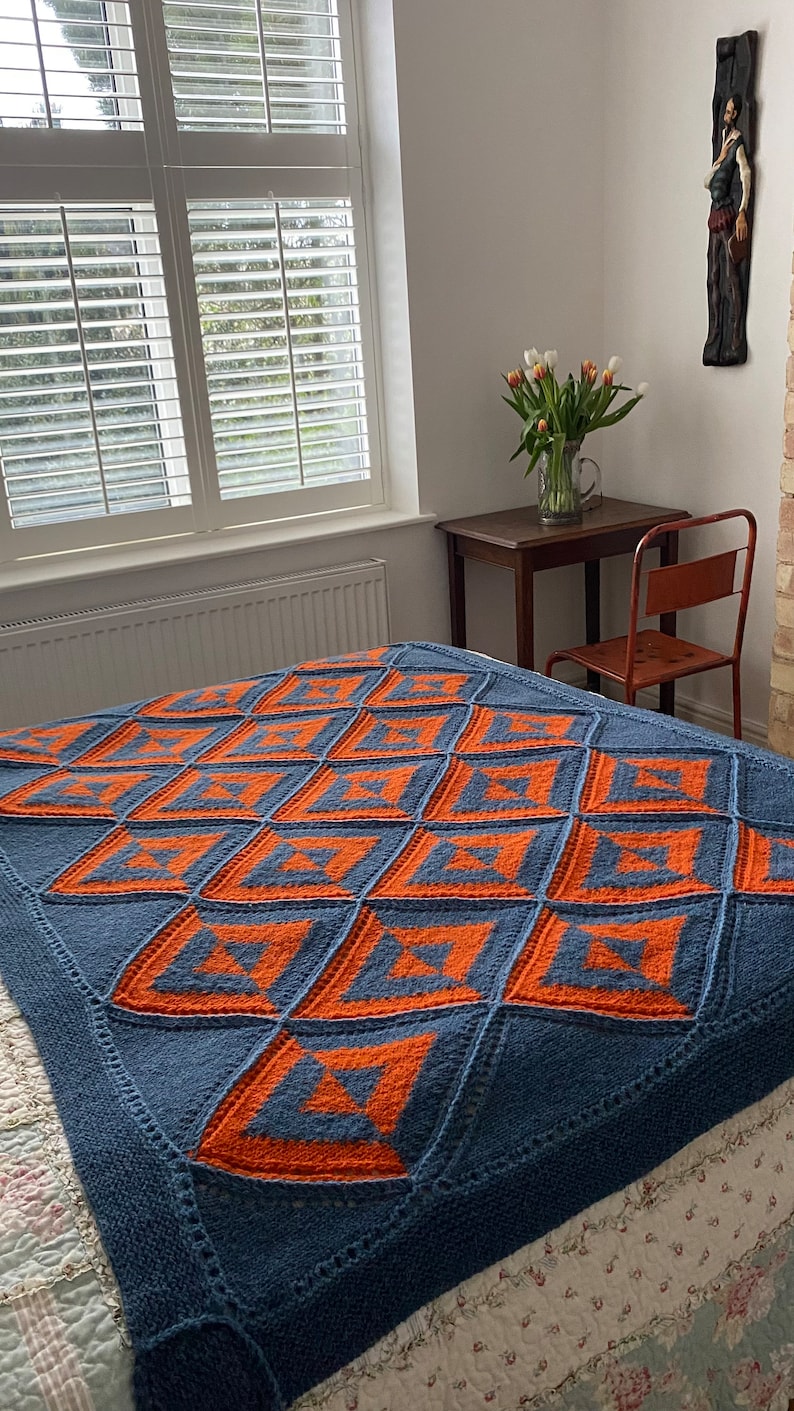 Pode incluir: Uma manta de malha azul e laranja com um padr&atilde;o geom&eacute;trico. A manta est&aacute; em uma cama com uma colcha branca e estampada com flores.