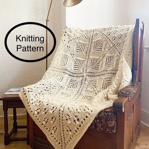 Può includere: Una coperta di lana bianca a maglia con un motivo traforato e pompon, drappeggiata su una poltrona in legno. Il testo "Knitting Pattern" è visibile nell'immagine.