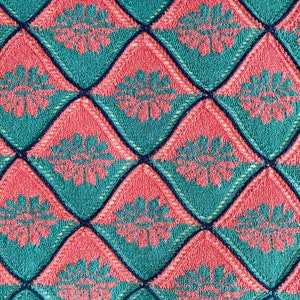 K&ouml;nnte beinhalten: Eine gestrickte Decke mit einem Rautenmuster in T&uuml;rkis und Rosa. Die Rauten haben ein florales Design in der Mitte.