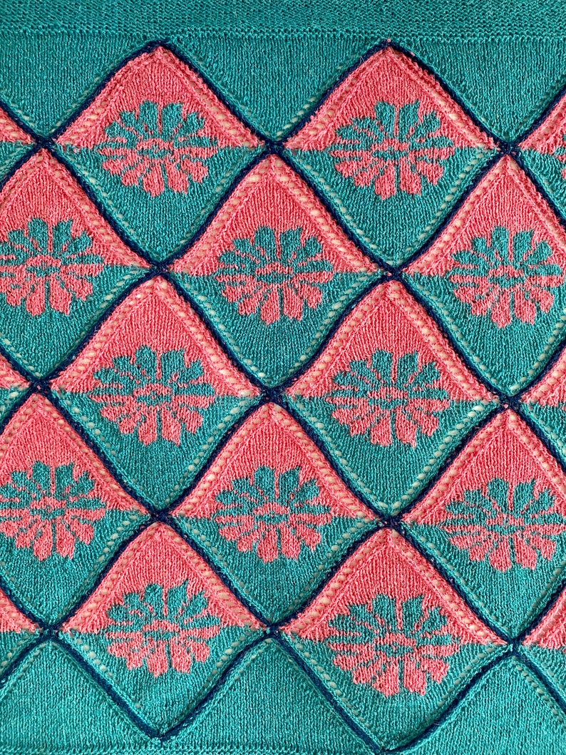 K&ouml;nnte beinhalten: Eine gestrickte Decke mit einem Rautenmuster in T&uuml;rkis und Rosa. Die Rauten haben ein florales Design in der Mitte.
