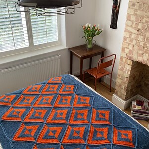 Pode incluir: Uma manta de malha azul e laranja com um padr&atilde;o geom&eacute;trico. A manta est&aacute; em uma cama em um quarto com uma parede branca e uma janela com persianas brancas.